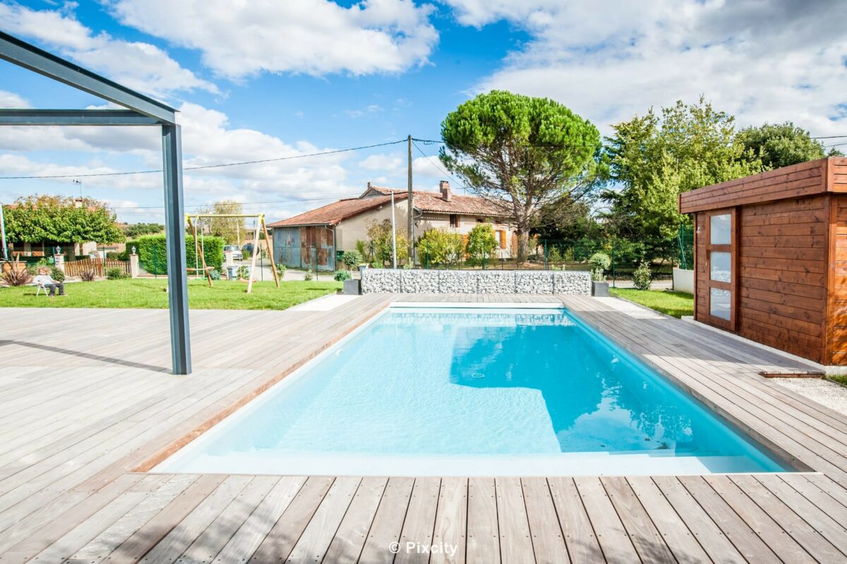 Aménagement terrasse bois autour piscine, piscine rectangulaire eau bleue claire, terrasse composite gris, pergola aluminium gris voile ombrage, abri jardin bois brun, maison grise fenêtres aluminium, pelouse verte, chaise jardin bleue, ciel nuageux dramatique