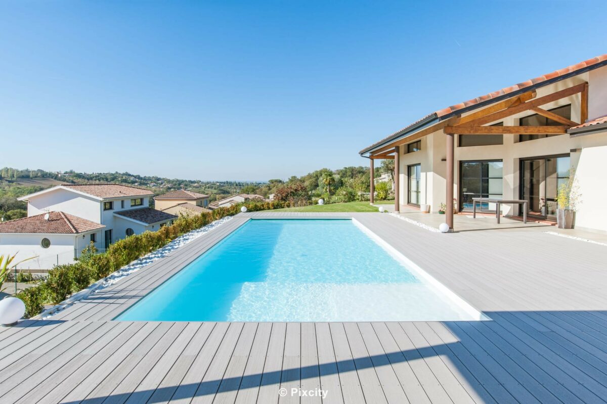 Aménagement terrasse bois autour piscine, très grande piscine rectangulaire longue eau cristalline, terrasse composite gris clair très spacieuse, villa luxe plain-pied baies vitrées, pergola bois poutres apparentes, pool house moderne, galets blancs, vue panoramique collines, ciel bleu immaculé