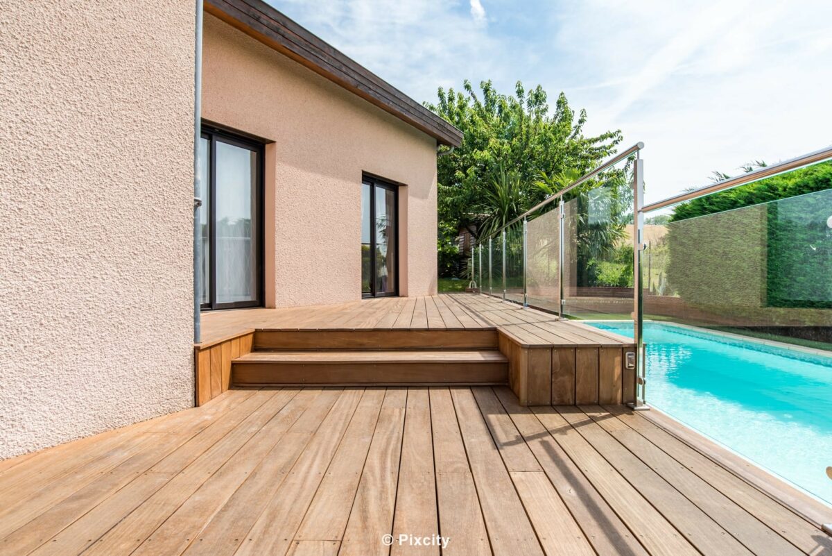 Aménagement terrasse bois autour piscine, petite terrasse bois exotique brun escalier marches, garde-corps inox et verre trempé, maison enduit beige baies vitrées aluminium noires, piscine bleue, palmier yucca, rosier, galets blancs, végétation luxuriante