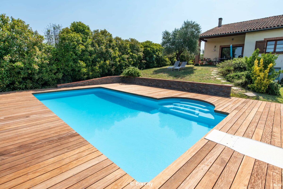 Aménagement terrasse bois autour piscine, piscine rectangulaire eau bleue, terrasse bois exotique brun chaleureux, olivier méditerranéen, pelouse verte, haie verte, maison provençale, ambiance champêtre estivale