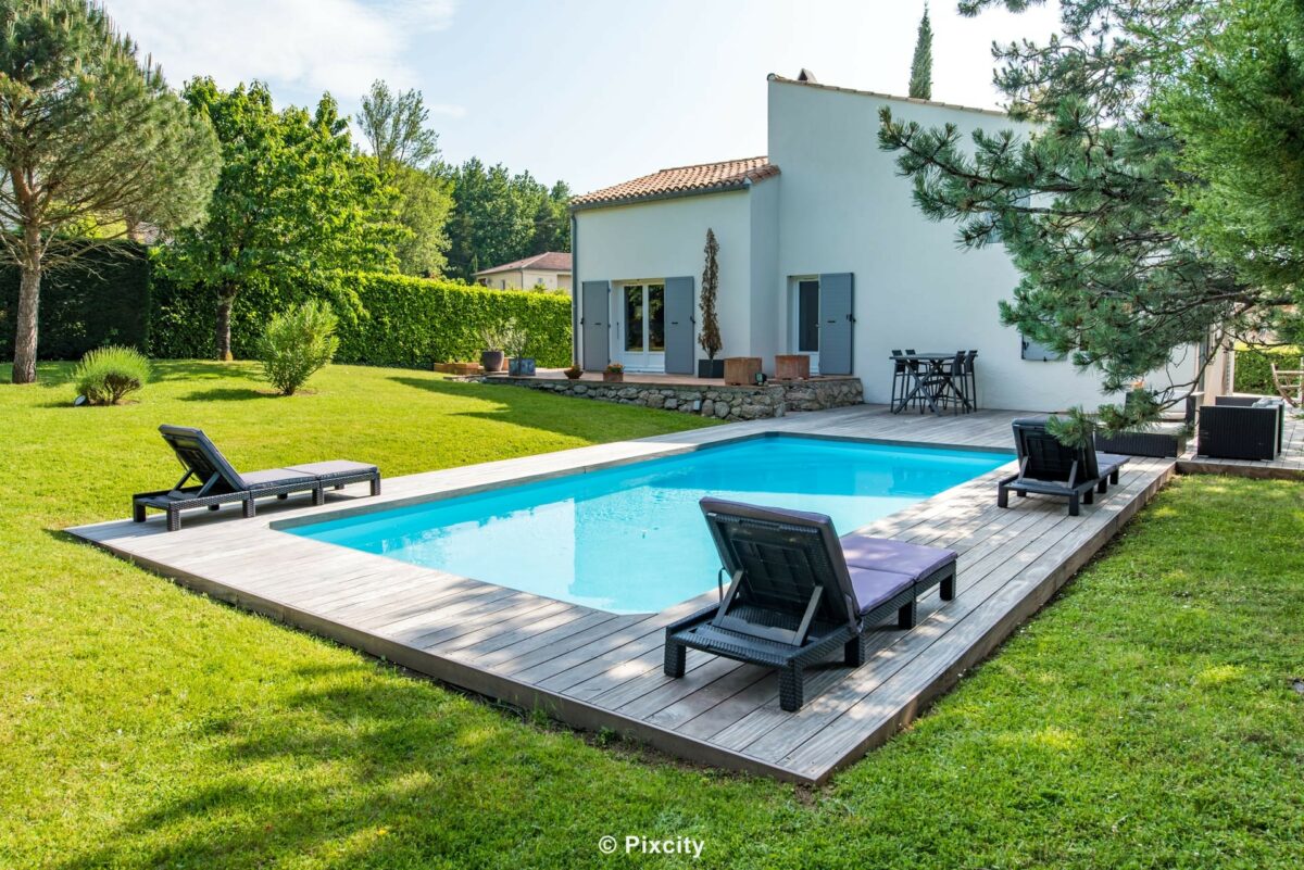 Aménagement terrasse bois autour piscine, très grande piscine longue eau cristalline, terrasse composite gris clair spacieuse, villa contemporaine provençale toiture plate, pool house chalet bois foncé, galets blancs, plantes méditerranéennes, ciel bleu vaste, vue quartier résidentiel