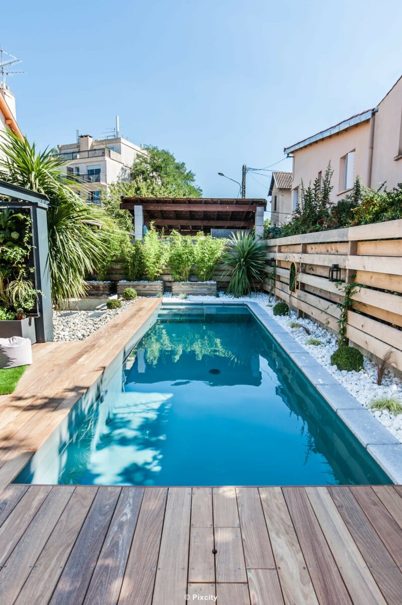 Aménagement terrasse bois autour piscine, piscine couloir longue eau sombre reflets, terrasse bois exotique clair, palmier trachycarpus, bambous, plantes tropicales exotiques, pergola bois suspendue, brise-vue lattes bois, pot rouge design, ciel bleu, jardin urbain