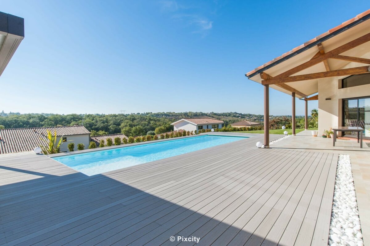 Aménagement terrasse bois autour piscine, grande terrasse composite gris très spacieuse, piscine longue rectangulaire eau bleue, villa pergola bois poteaux apparents, vue panoramique dégagée collines quartier résidentiel, ciel bleu vaste, galets blancs, jardin soigné