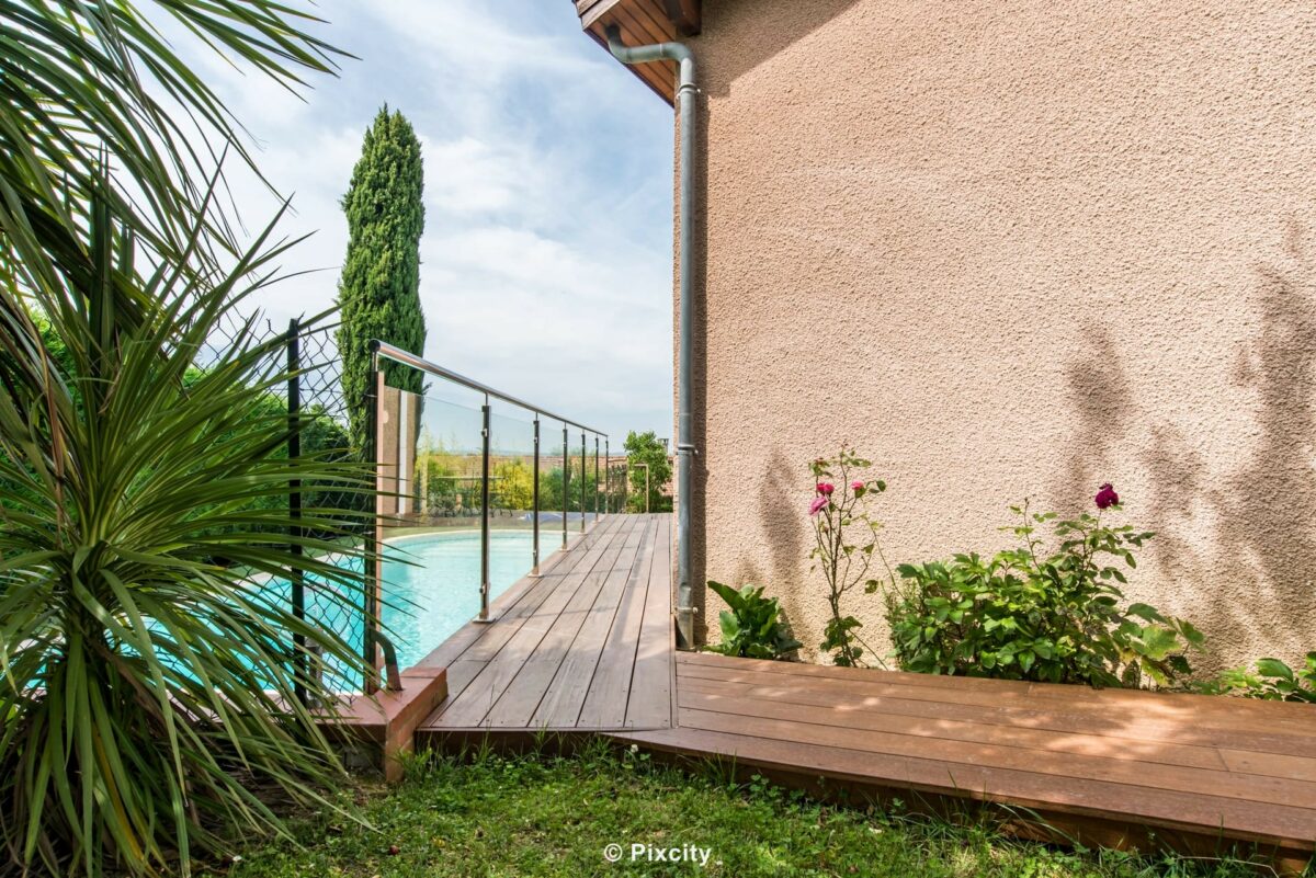 Aménagement terrasse bois autour piscine, terrasse bois exotique brun étroite avec marches, palmier yucca, garde-corps inox et verre transparent, piscine bleue fond, cyprès méditerranéen, rose en fleur, maison enduit, végétation printemps