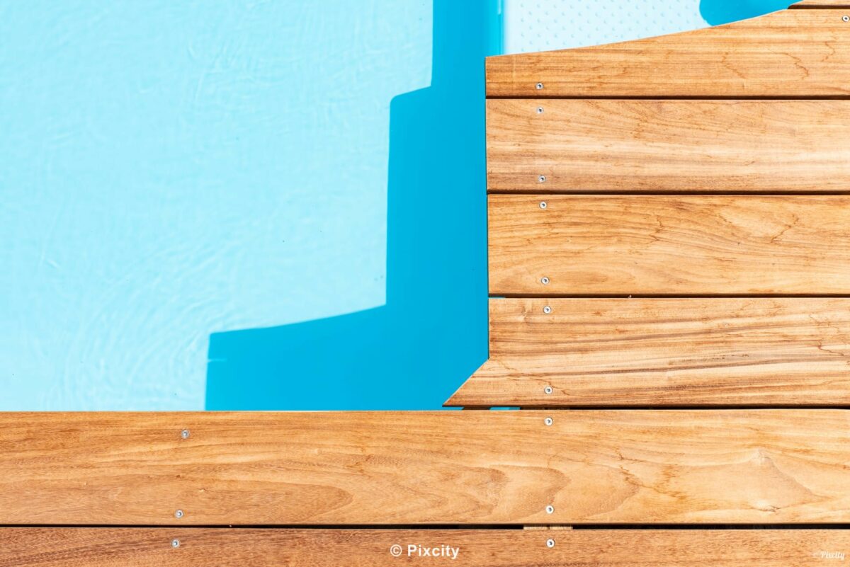 Aménagement terrasse bois autour piscine, gros plan jonction terrasse bois exotique brun doré et piscine eau bleu turquoise, escalier immergé en L marches blanches, ombres graphiques géométriques, jeux de lumière, texture bois foncé, minimaliste