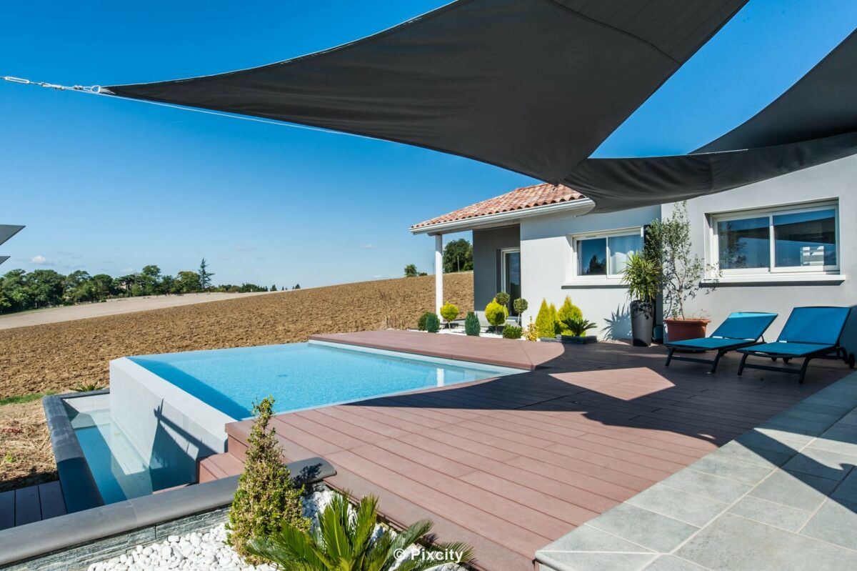 Aménagement terrasse bois autour piscine, terrasse composite brun rouge, piscine avec bassin bain de pieds débordement latéral, voiles d'ombrage triangulaires noires tendues, villa moderne enduit blanc toits tuiles, chaises longues bleues, yucca agave oliviers, pelouse, campagne fond, ciel bleu