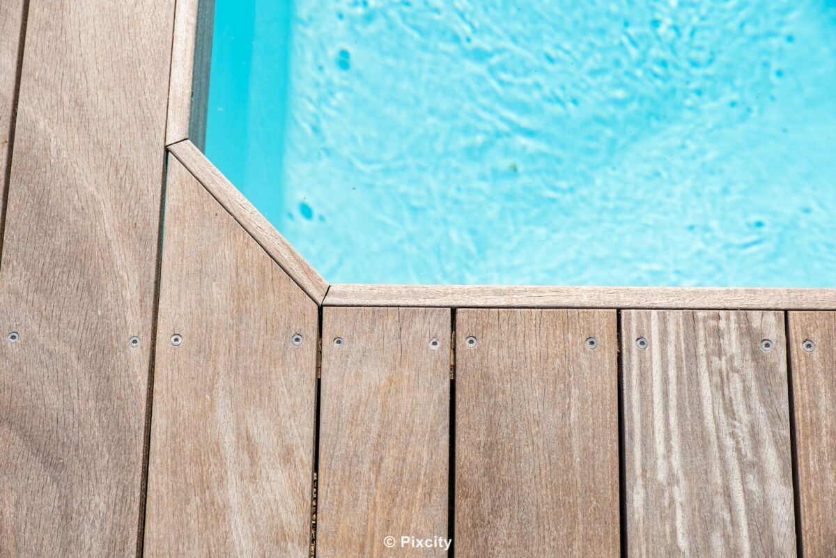 Aménagement terrasse bois autour piscine, gros plan coin angle terrasse bois gris patiné et eau piscine turquoise claire, jonction nette bois eau, texture bois horizontal et reflets eau lumière, composition minimaliste macro