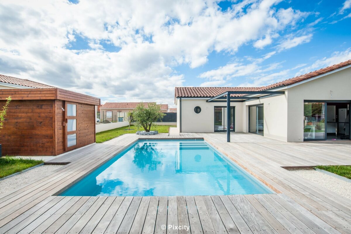 Aménagement terrasse bois autour piscine, grande terrasse composite gris, piscine rectangulaire eau bleue, villa plain-pied enduit blanc moderne, pergola aluminium noir baies noires, abri jardin bois brun foncé, pelouse, galets, olivier, ciel nuageux dramatique blanc