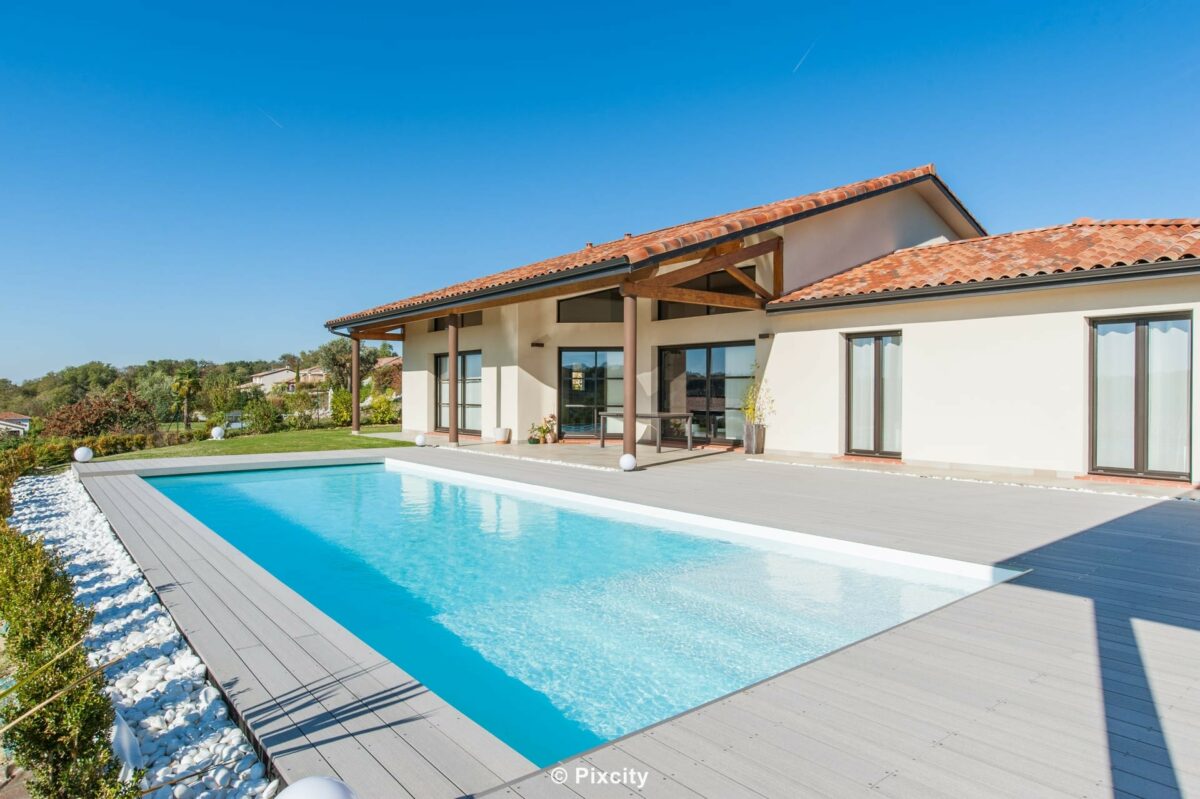 Aménagement terrasse bois autour piscine, grande piscine rectangulaire bleue, terrasse bois composite gris, villa contemporaine toits tuiles, pergola bois, pool house baies noires, galets blancs, plantes méditerranéennes yucca, ciel bleu, lumière dorée