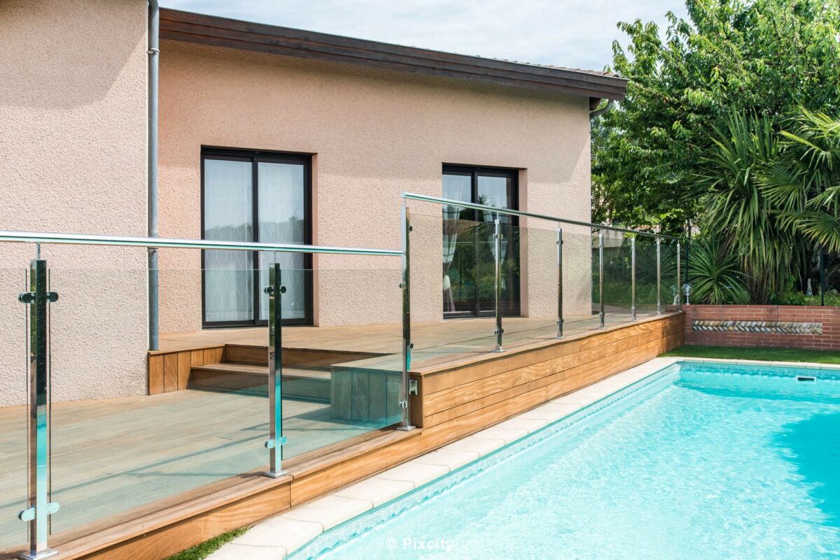 Aménagement terrasse bois autour piscine, piscine rectangulaire bleue, garde-corps inox et verre trempé sur terrasse bois exotique, maison enduit beige fenêtres aluminium noires, palmier yucca, rosier fleurs roses, végétation jardin méditerranéen