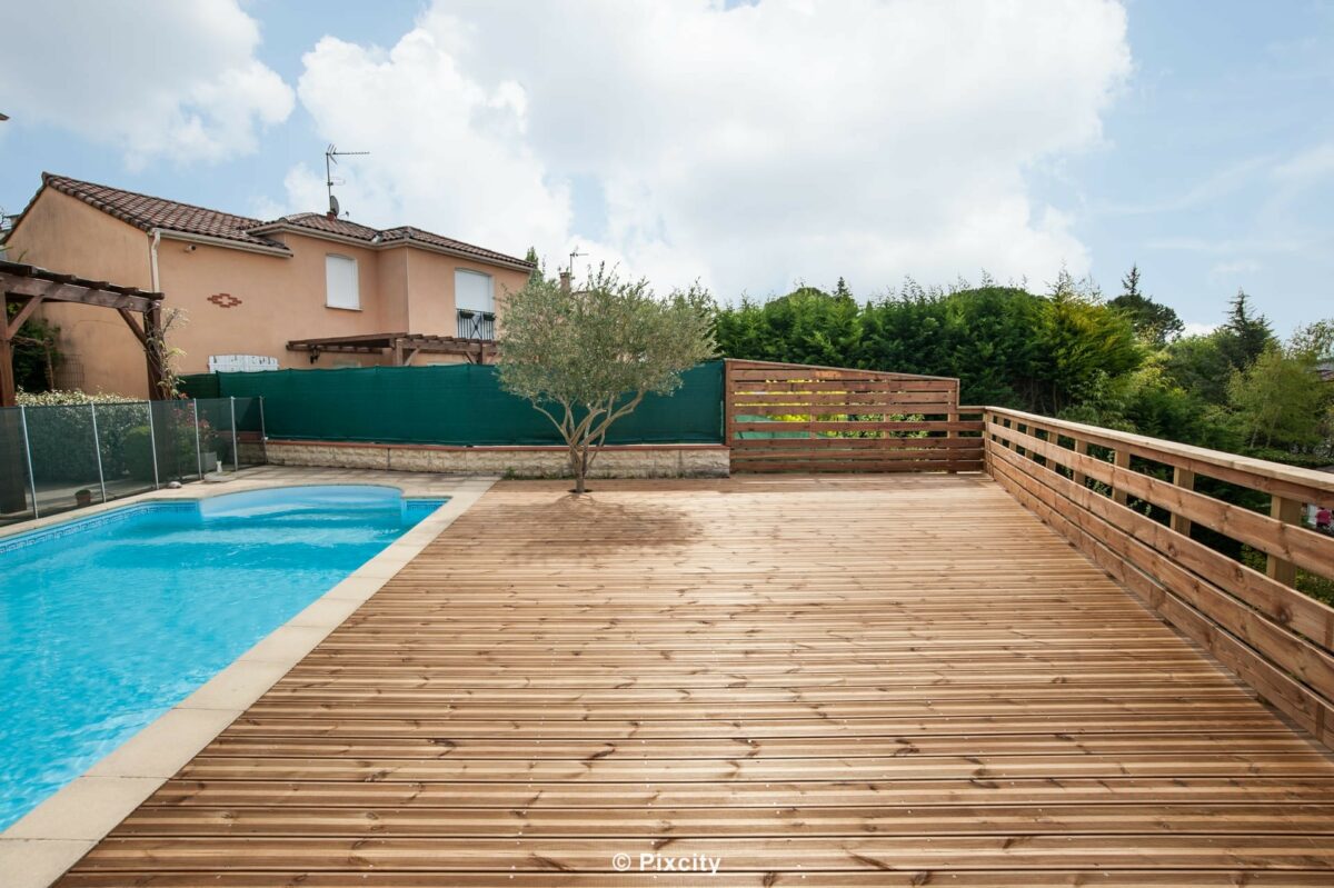 Aménagement terrasse bois autour piscine, grande terrasse bois pin brut clair très spacieuse, olivier méditerranéen planté dans orifice terrasse, palissade bois horizontal brun, garde-corps lattes horizontales bois, piscine bleue côté, pelouse verte, ciel gris doux