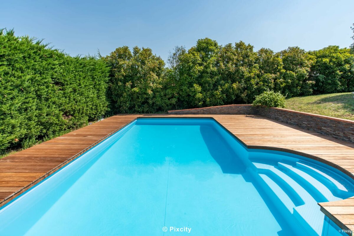 Aménagement terrasse bois autour piscine, piscine rectangulaire eau turquoise fraîche, terrasse bois exotique teck brun doré, haie thuyas denses bien taillés, pelouse verte, pot céramique artisanal bleu pétrole, ombres légères, lumière chaude été campagne